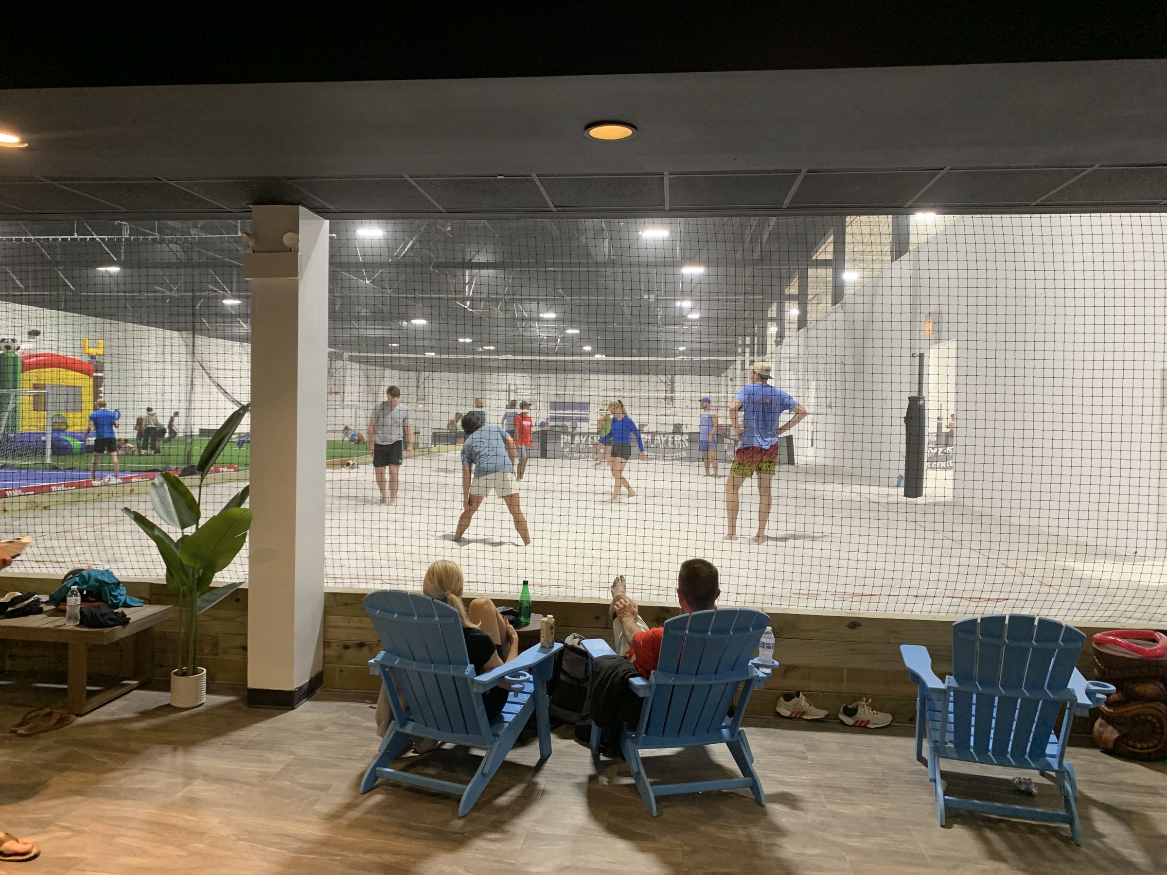 Players Beach lounge at Players Sports Center — blue Adirondack chairs facing a full indoor sand volleyball court, with turf and a bounce house in the background