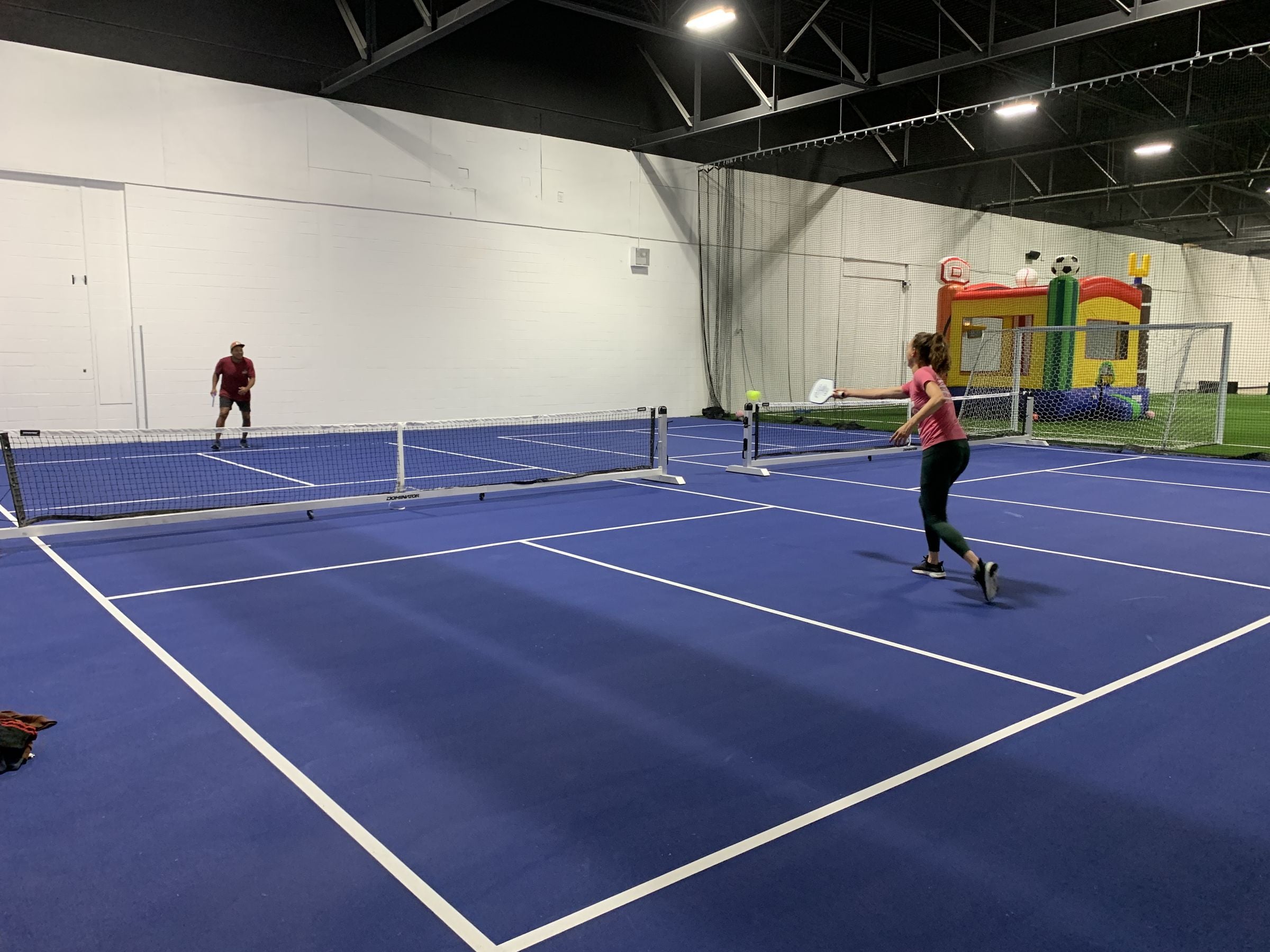 Pickleball rally on the blue indoor courts at Players Sports Center, with the turf field in the background